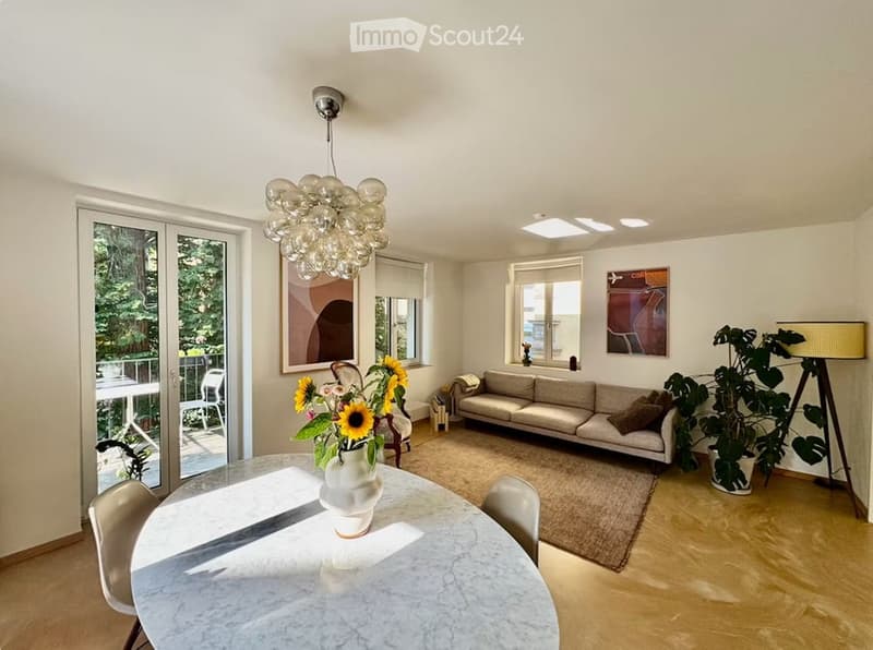 Bright living and dining area with a modern sofa, dining table with chairs, a large chandelier, and a view of a balcony.