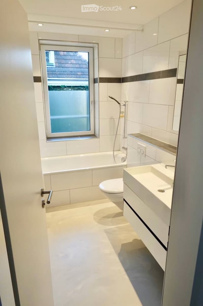 Modern bathroom with white tiles, a bathtub, toilet, sink, shower, and a window providing natural light.