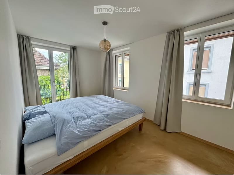 Bright bedroom with wooden flooring, large windows with curtains, a single bed with blue bedding, and a hanging light fixture.