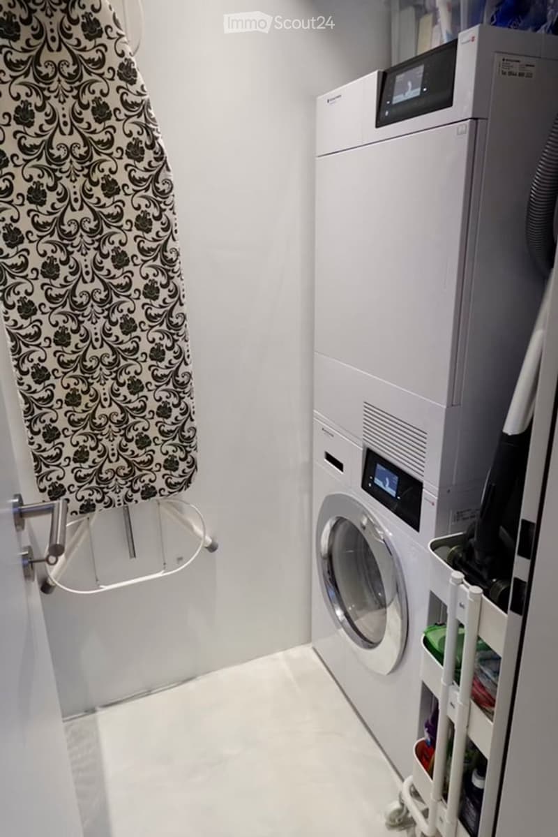 Modern bathroom with white tiled floor and walls, featuring a washing machine, dryer, and an ironing board with a patterned cover.