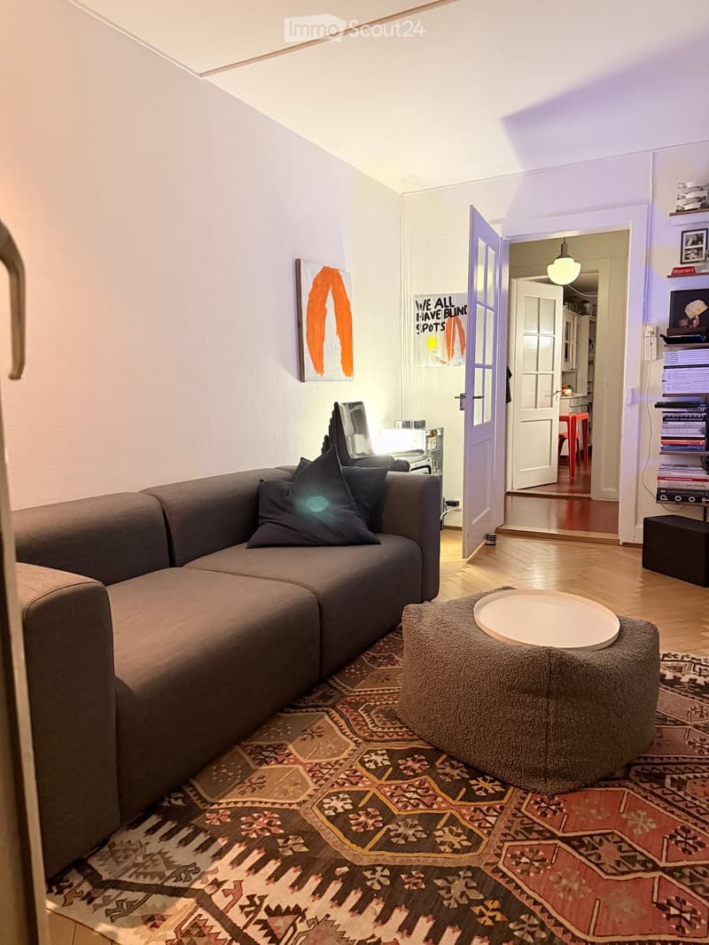 Living room with a grey sectional sofa, ottoman, round table, shelves, and artwork on the wall. The room features wooden flooring and a patterned rug.