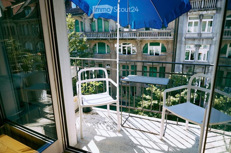 Balcony with two white chairs, a small table, and a blue umbrella, overlooking a historic building with green shutters.