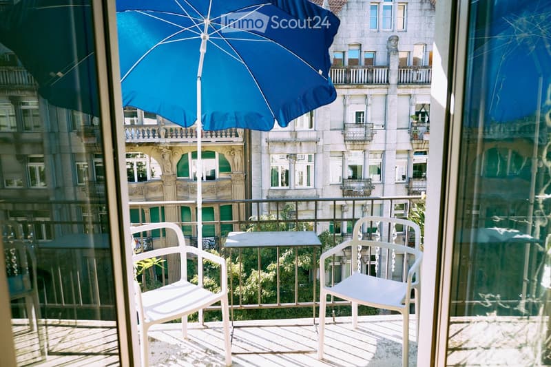 Balcony with two white chairs, a small table, and a large blue umbrella, overlooking a cityscape with historic buildings and greenery.