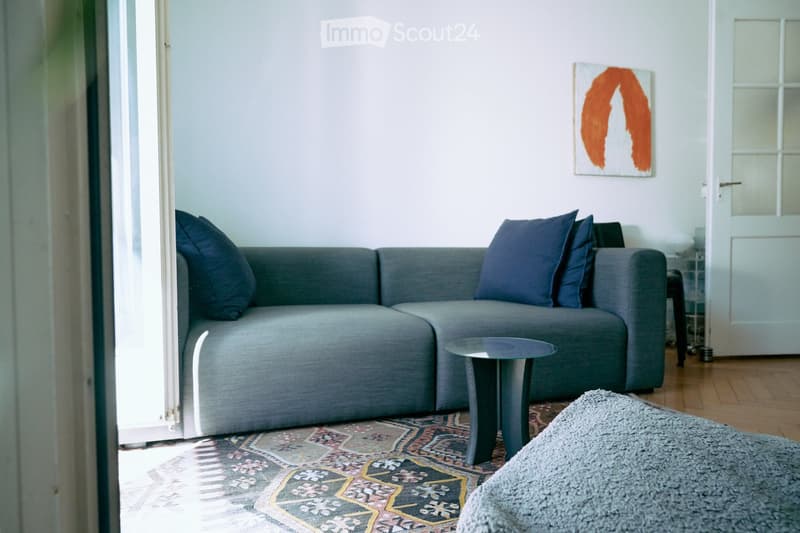 Modern living room with a grey sectional sofa, blue throw pillows, a small round coffee table, a patterned rug, and a contemporary painting on the wall.