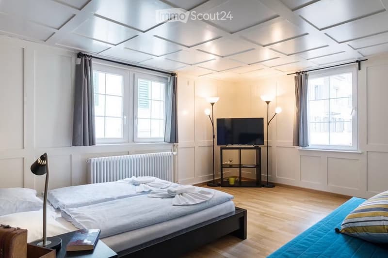 Cozy, well-lit bedroom with two single beds, a nightstand with a lamp, a small table with a television, and a chair. The room features wooden flooring, white walls, and large windows with curtains.