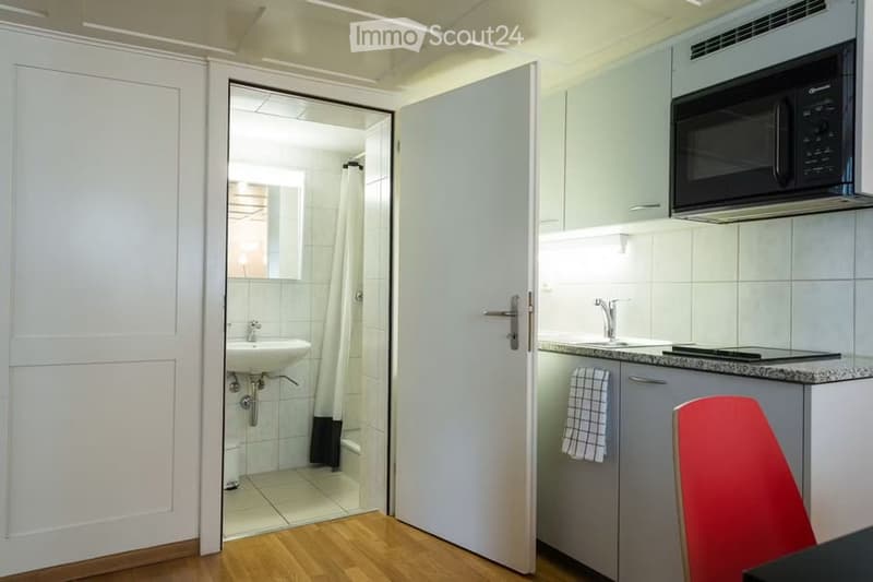 A small, modern kitchen with a microwave, sink, and faucet, connected to a compact bathroom with a shower and sink. A red chair is visible near the kitchen counter.