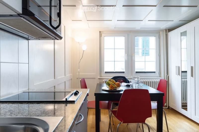 A modern kitchen with a dining area, featuring a black dining table with red chairs, a bowl of fruit, and a sleek kitchen counter with an induction cooktop and built-in oven.