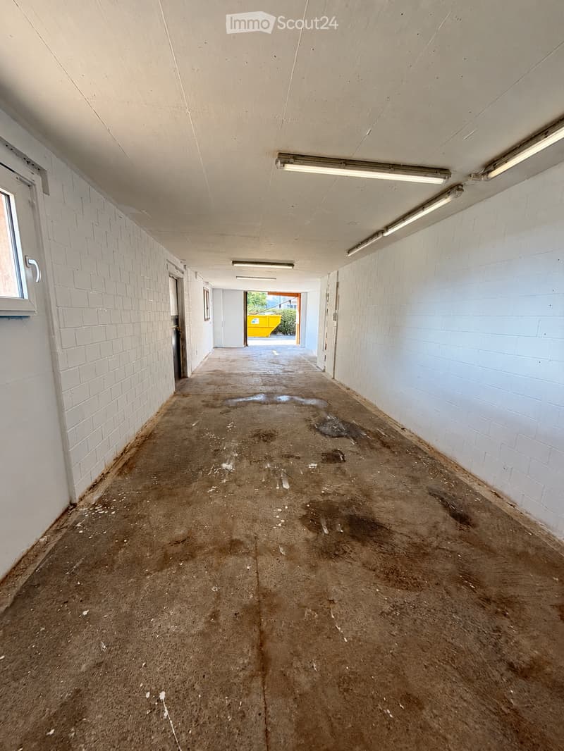 Empty corridor with white brick walls, concrete floor, and fluorescent lighting. The floor shows signs of wear and dirt, with a small puddle visible.