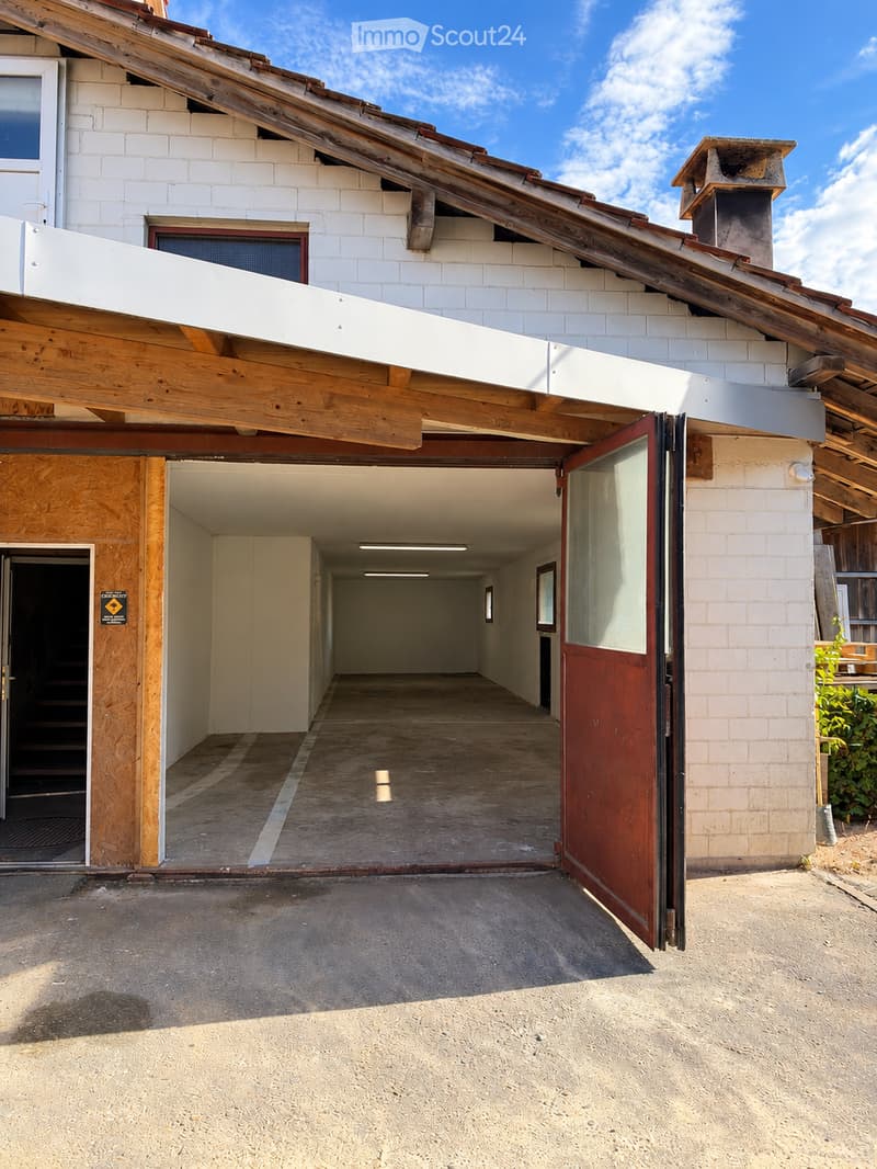 A modern building with a large garage entrance, white brick walls, wooden beams, and a slanted wooden roof. The garage has a concrete floor and is partially open, revealing a hallway inside.