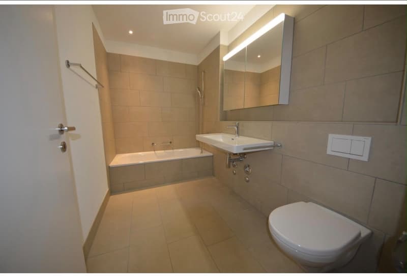 Modern bathroom with beige tiles, a bathtub, a sink, a toilet, and a large mirror with built-in lighting.