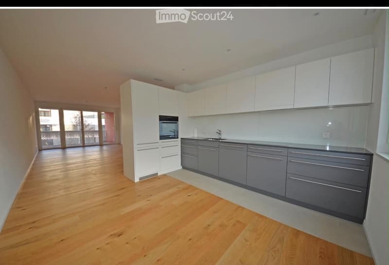 Modern kitchen with white and grey cabinets, wooden flooring, and large windows leading to a balcony.