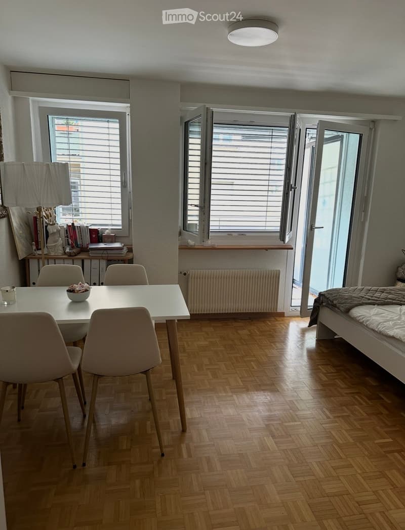 Bright, open room with wooden flooring, large windows, a dining table with chairs, a bed, a bookshelf, and a lamp.