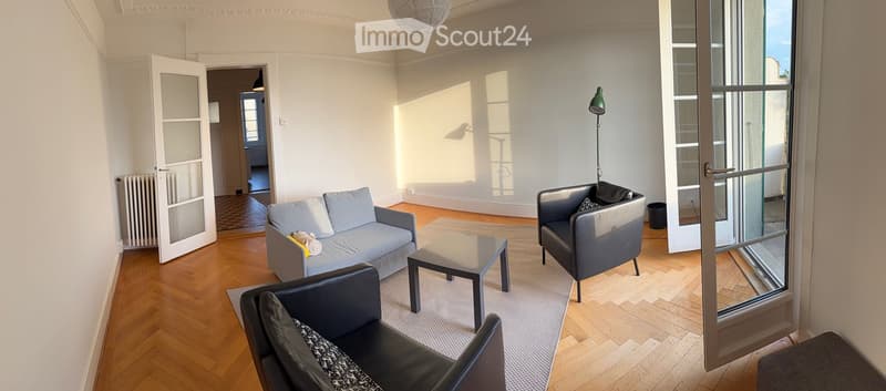 Bright living room with wooden flooring, large windows, and modern furniture including a sofa, coffee table, and armchairs.