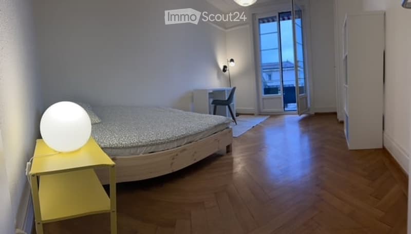 Bright bedroom with wooden flooring, a double bed with patterned bedding, a yellow nightstand with a round lamp, a desk with a chair, and large windows with a view.