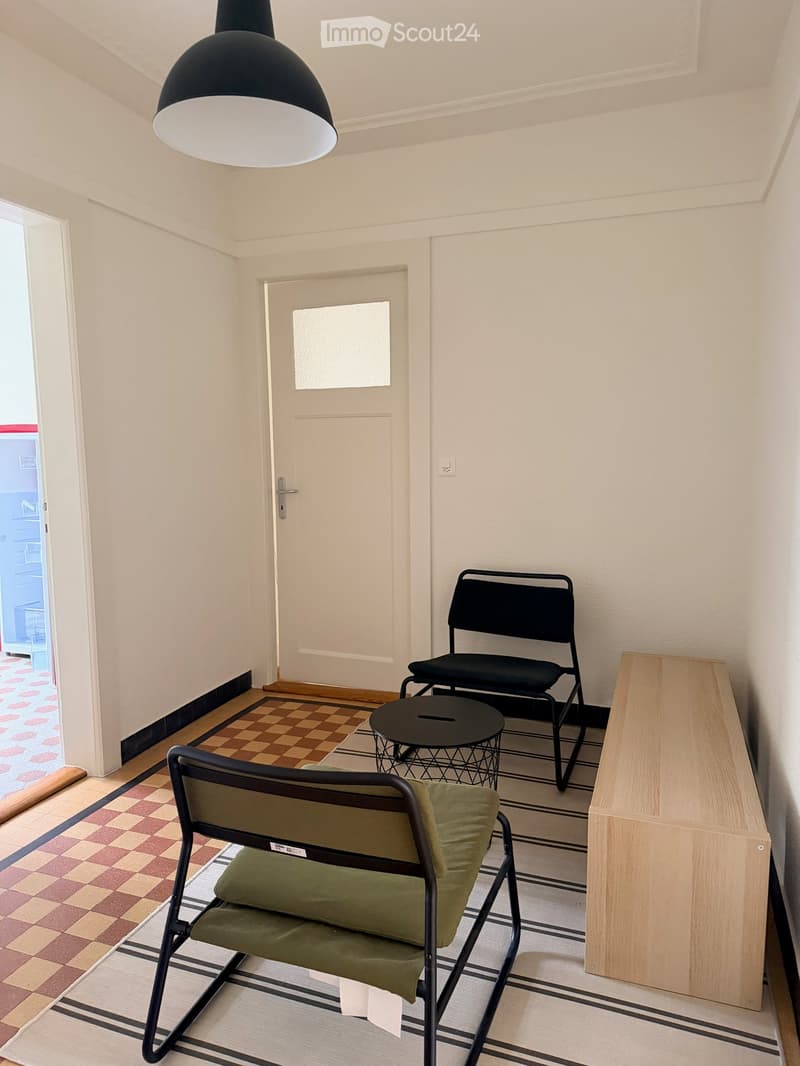 A small, furnished room with a wooden floor, a black hanging lamp, a green chair, a black chair, a small round table, and a wooden cabinet.
