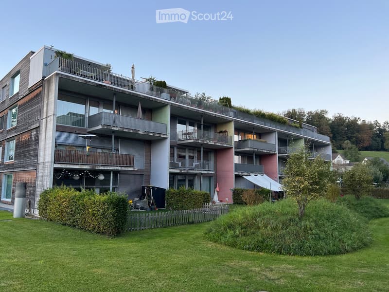 Modern multi-story residential building with balconies, greenery on the roof, and a well-maintained lawn in front.