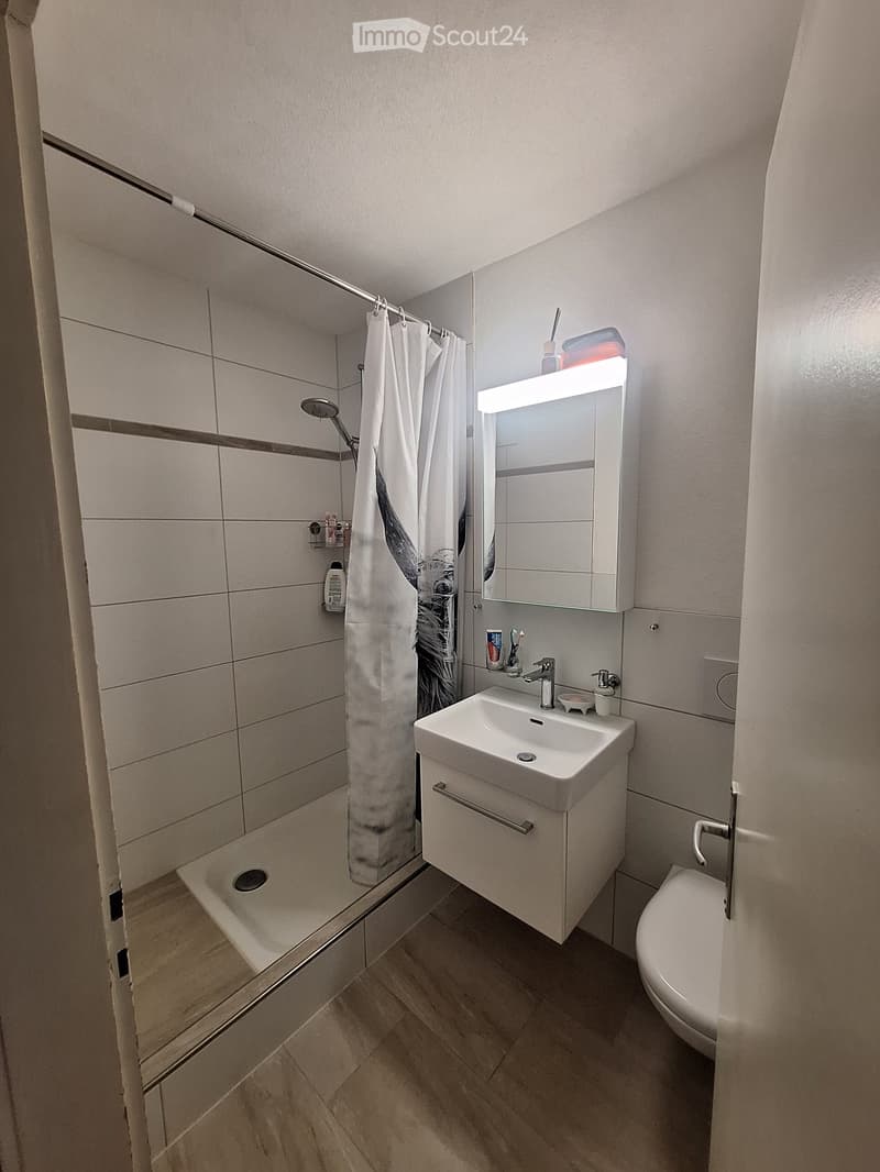 Modern bathroom with a white sink, mirror, toilet, and shower area with a curtain. The floor is tiled, and the walls are tiled with a neutral color scheme.