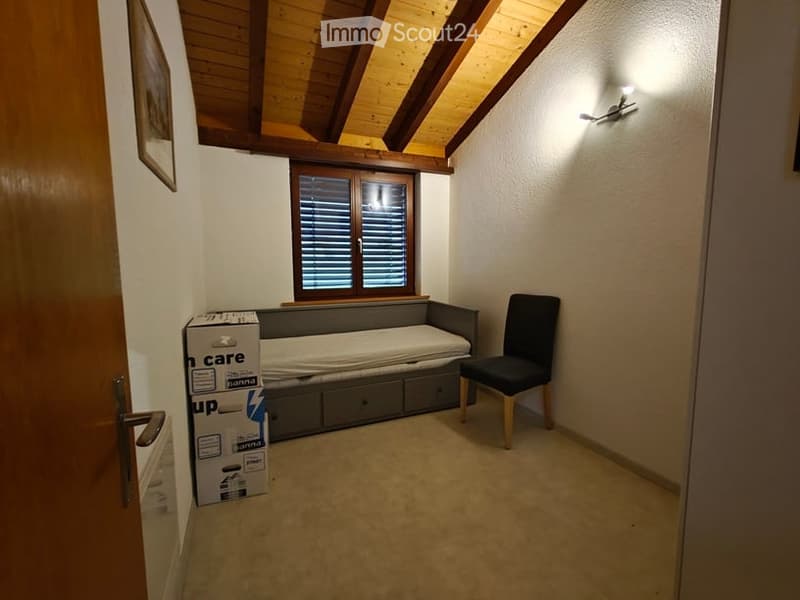 Cozy attic bedroom with wooden slanted ceilings, a window with shutters, a sofa bed, a chair, and a storage unit. A cardboard box is placed near the door.