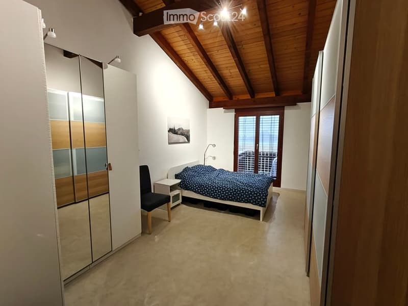 Cozy bedroom with a slanted wooden ceiling, large window offering a view, a bed with blue bedding, a chair, a nightstand, and a modern wardrobe with mirrored doors.