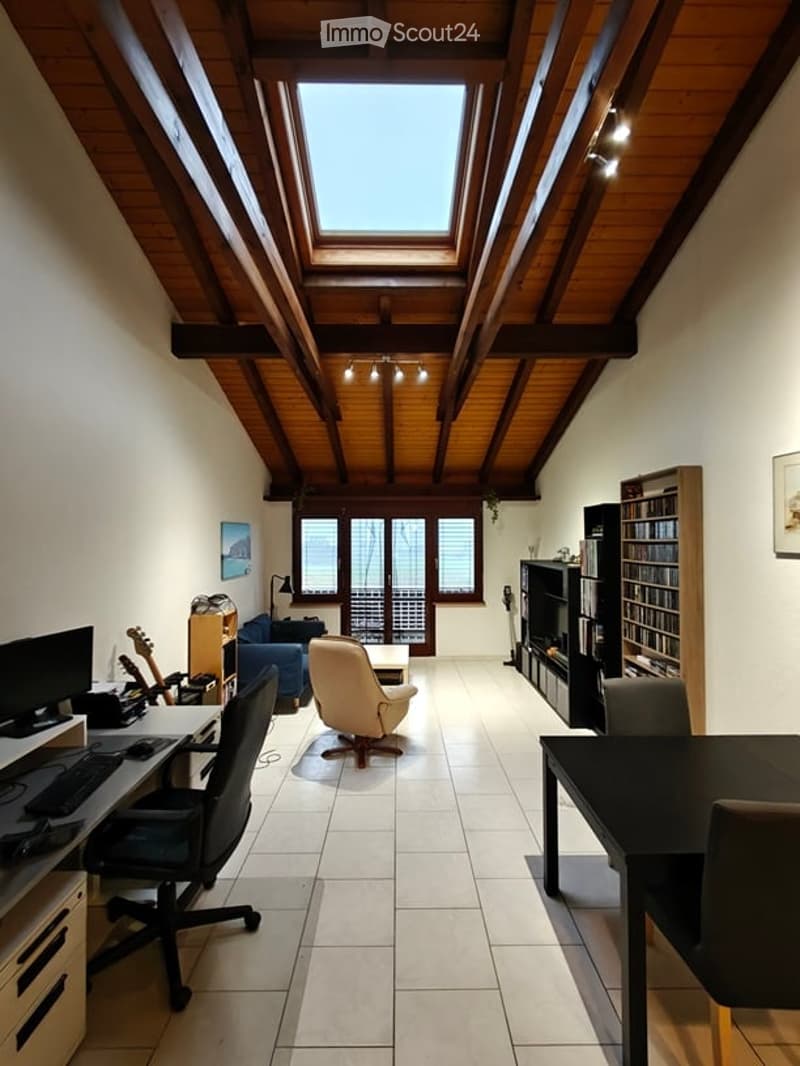 Bright, multi-functional room with wooden slanted ceiling, skylight, and large windows offering a view. Contains a desk with a computer, a comfortable armchair, a sofa, a dining table, and a bookshelf.