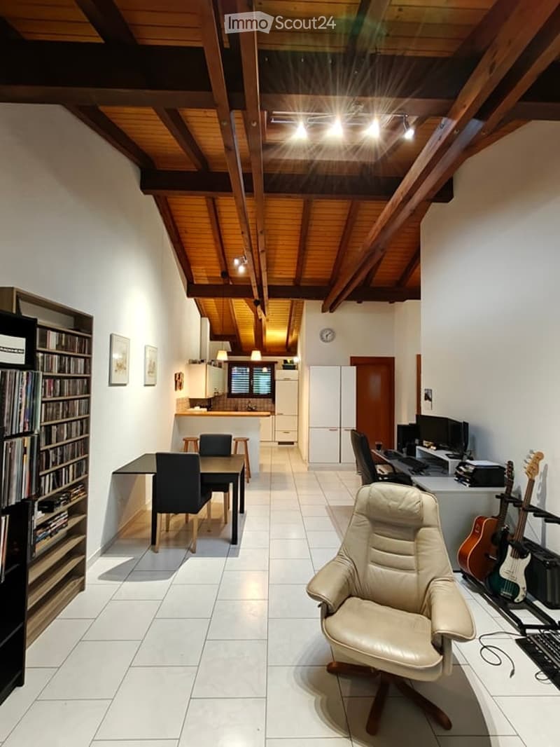 A modern, open-plan living and dining area with exposed wooden beams on the ceiling, white tiled flooring, and a mix of contemporary and rustic furniture. The space includes a dining table with chairs, a comfortable leather armchair, a bookshelf, a kitche