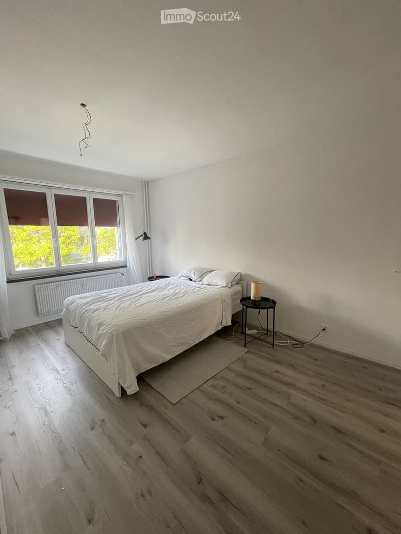 Minimalist bedroom with a white bed, nightstand, lamp, and small table near a large window with blinds.