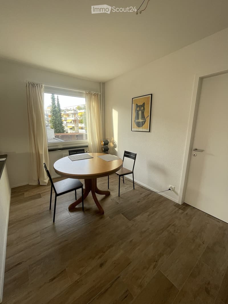 Dining area with wooden floor, round wooden table, four chairs, and a framed picture on the wall. Large window with curtains offers a view of the outside.