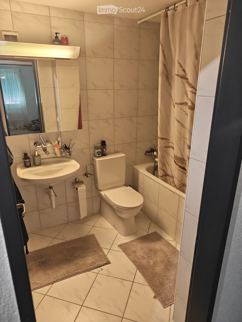 Modern bathroom with beige tiles, a sink, toilet, bathtub, and shower curtain. Includes a mirror, toiletries, and two rugs on the floor.