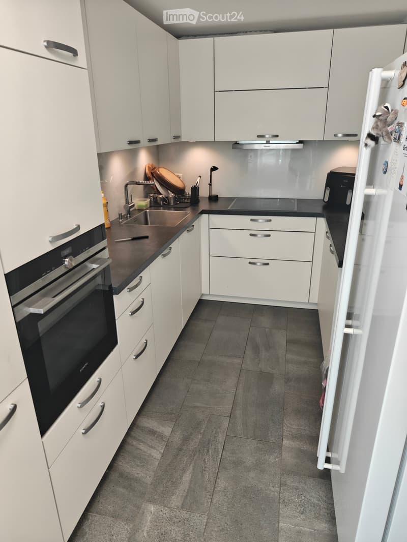Modern kitchen with white cabinets, stainless steel appliances, and a dark countertop. Features include an oven, refrigerator, sink, and various kitchen utensils.