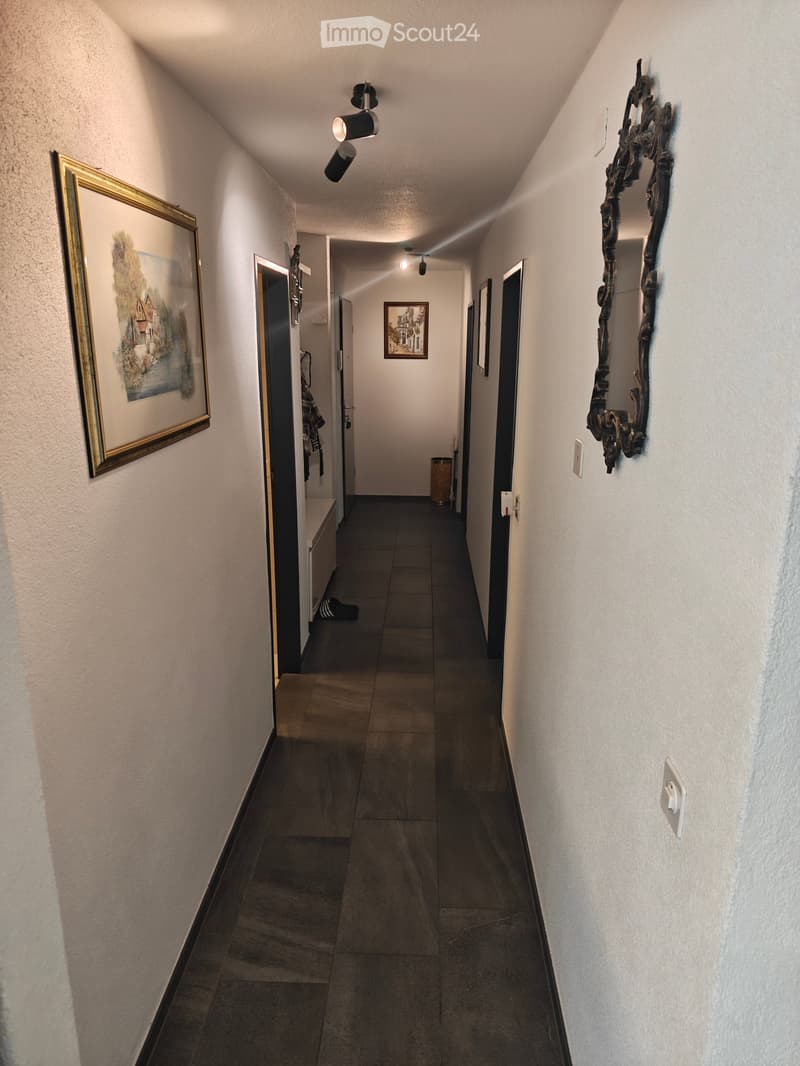 A narrow hallway with tiled flooring, white walls, and several doors leading to different rooms. There are framed pictures on the walls and a small bench with a shoe placed on it.