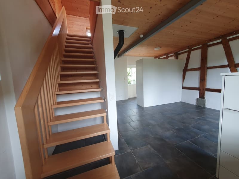 Wooden staircase with a handrail leads to an upper level. The hallway has dark tiled flooring, white walls, and exposed wooden beams on the ceiling.