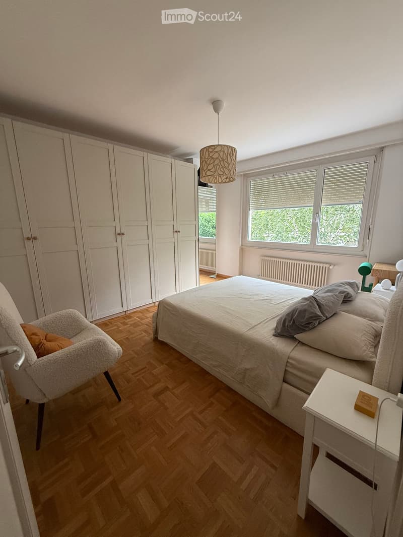 Modern bedroom with wooden flooring, large bed, nightstand, armchair, and built-in wardrobe.