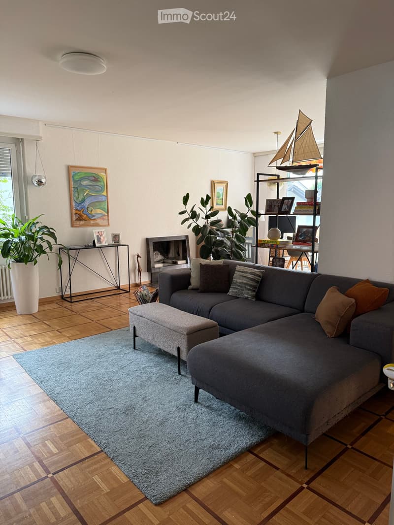 Modern living room with a grey sectional sofa, ottoman, coffee table, fireplace, and decorative shelf with books and a model sailboat.