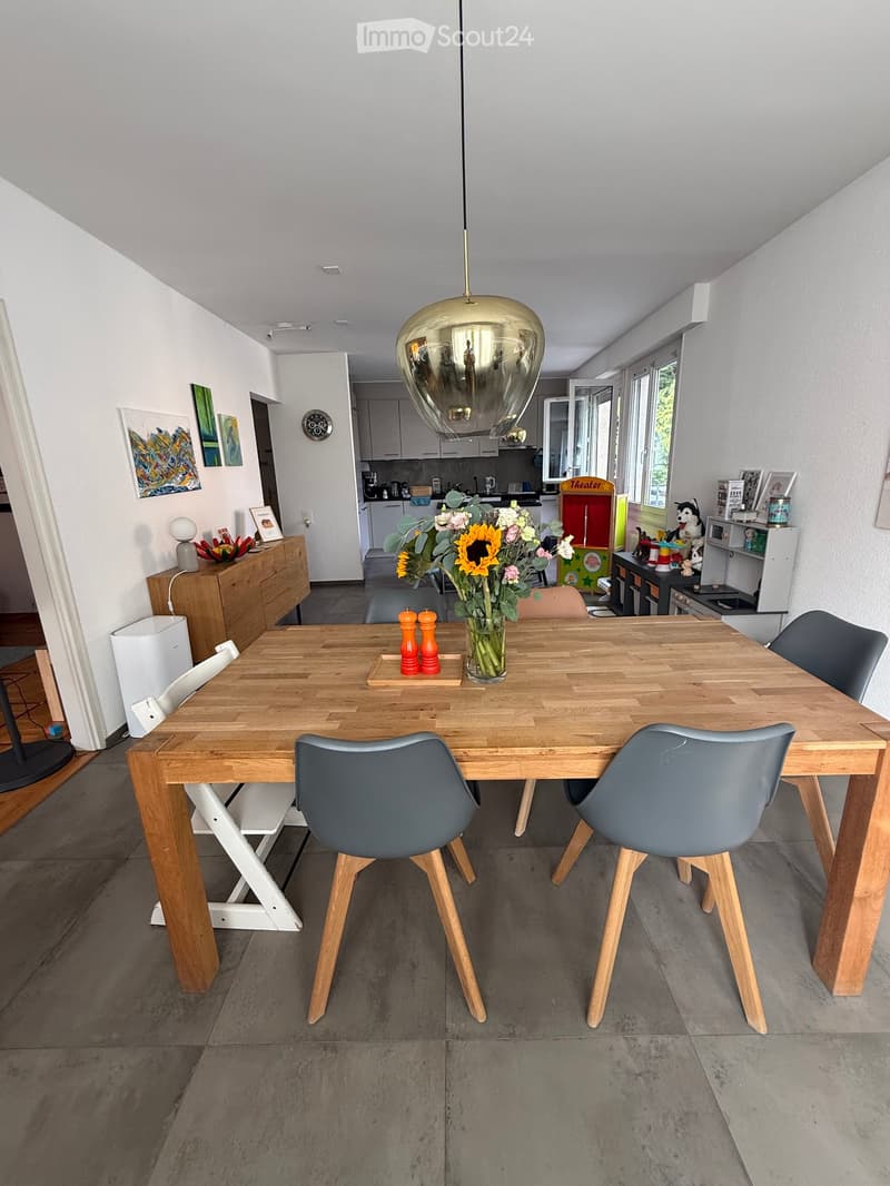 The image shows a modern kitchen and dining area with a wooden dining table and chairs, a vase with flowers, and a kitchen area with cabinets, appliances, and a light fixture.