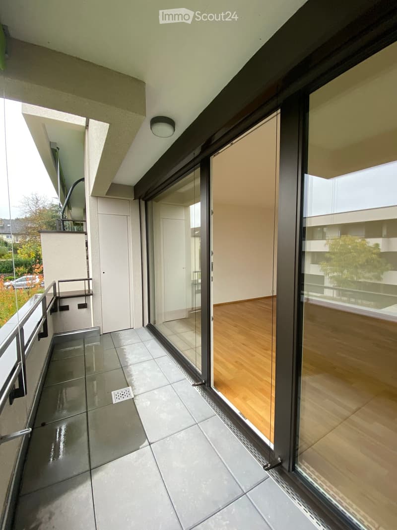 Modern balcony with tiled floor, glass sliding doors, metal railing, and a view of the surrounding area.