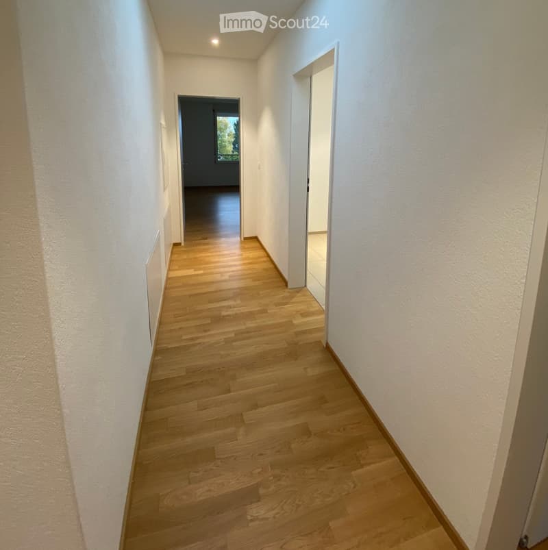 A narrow hallway with light wooden flooring and white walls, featuring two doors at the end and a small window letting in natural light.