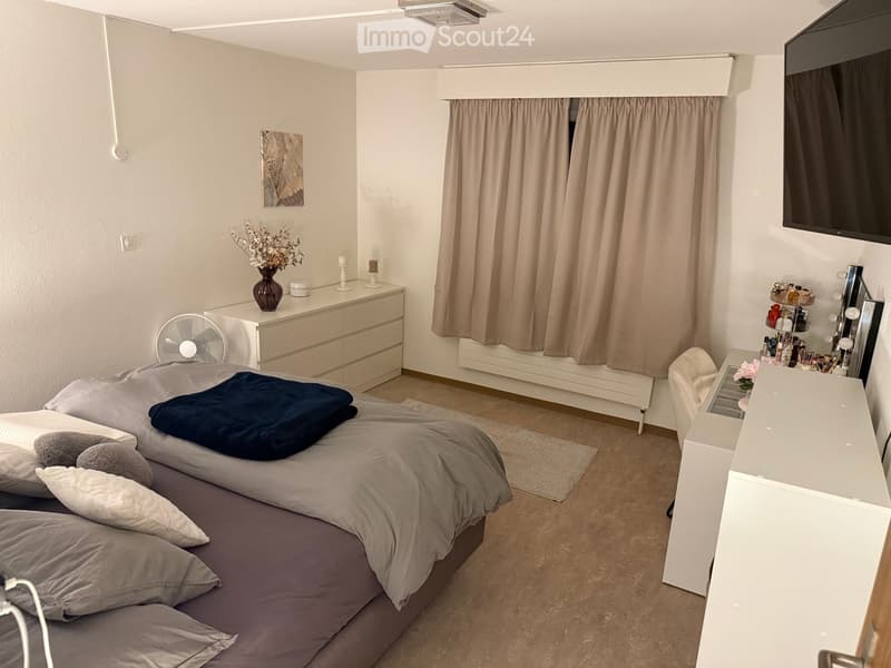 Modern bedroom with a double bed, nightstand, mirror, fan, vase with flowers, and a TV mounted on the wall.
