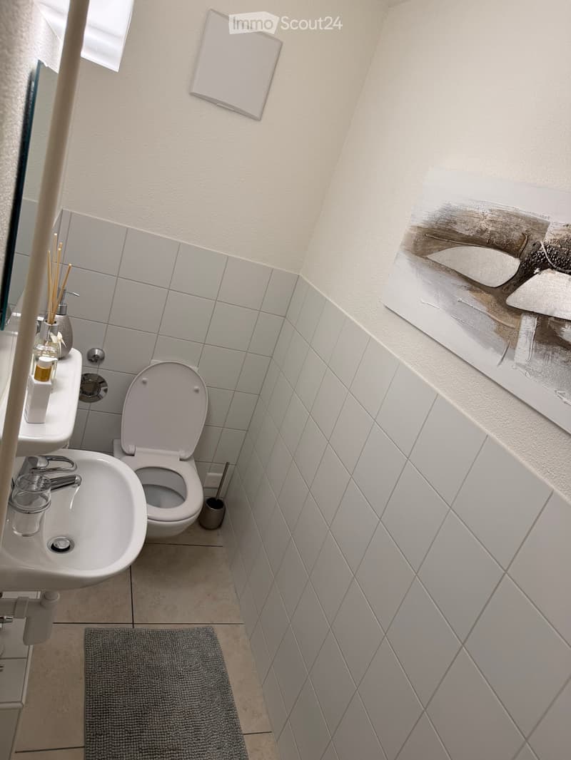 A compact bathroom with a toilet, sink, and mirror. The walls are tiled in a light grey color, and a small grey rug is placed on the floor.