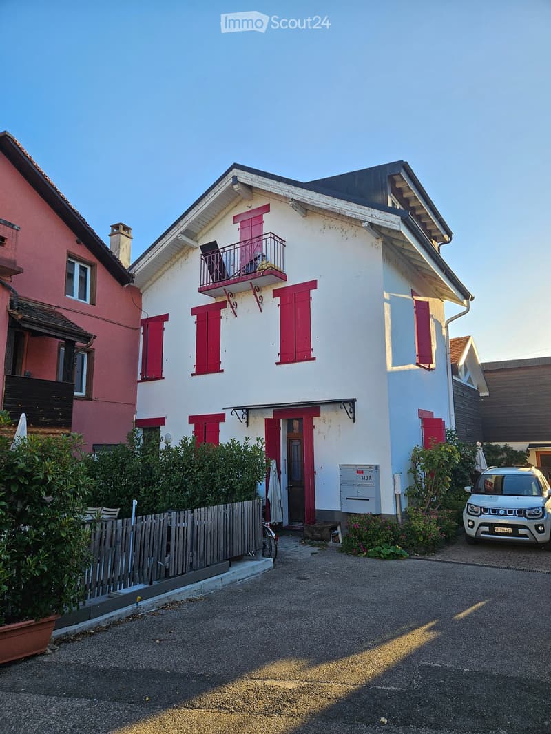 Two-story house with white walls, red shutters, and a small balcony on the upper floor. The house has a wooden fence and is surrounded by greenery and a parked car.