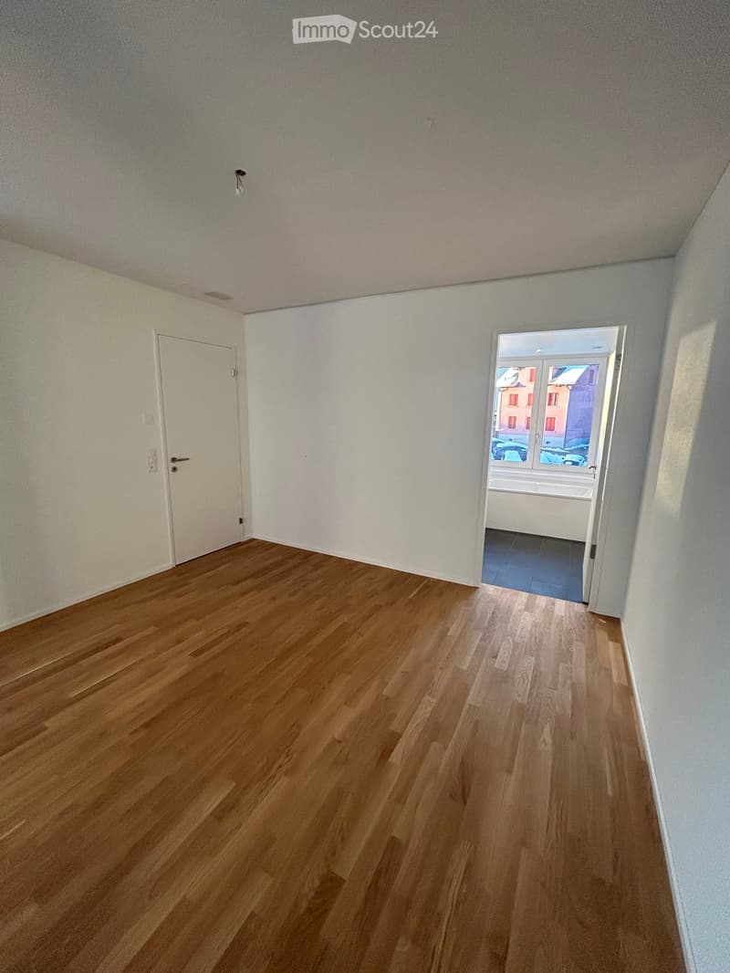 Empty room with wooden flooring, white walls, and a door leading to another room.