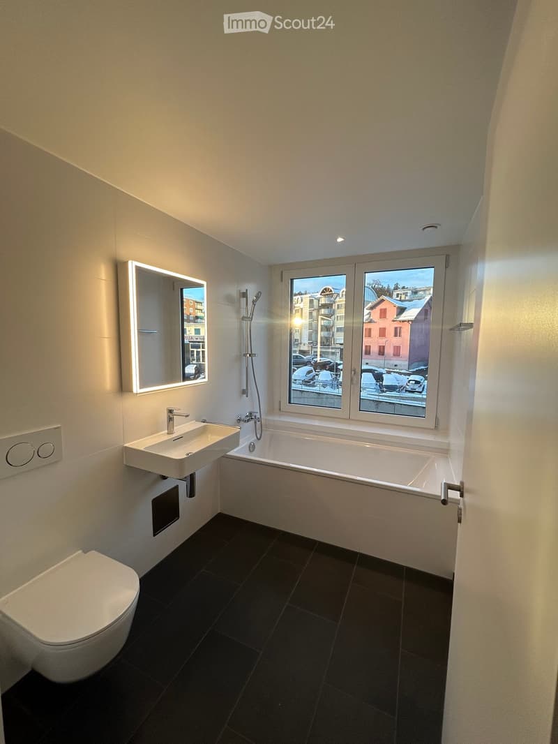 Modern bathroom with a white toilet, sink, bathtub, and illuminated mirror. The room features dark tiled flooring and a large window offering a view of the street.