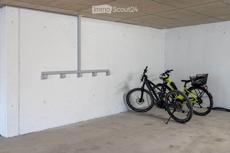 Two bicycles, one black and one green, are parked in a concrete-walled area with a metal bike rack attached to the wall.