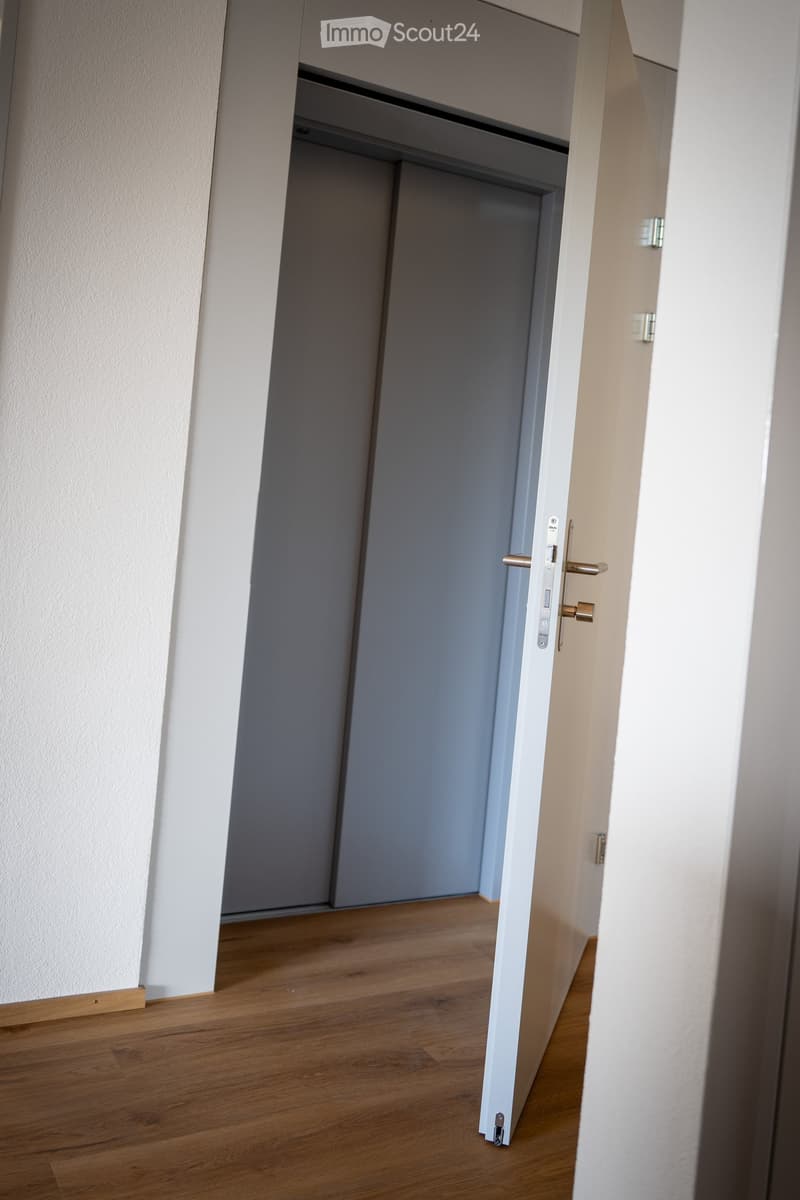 A narrow hallway with light wooden flooring and white walls, featuring an open door leading to another room.