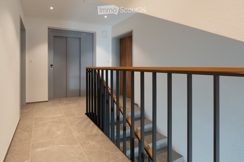 Modern hallway with tiled floor, white walls, and a staircase with wooden steps and metal railing. Two doors are visible, one grey and one wooden.
