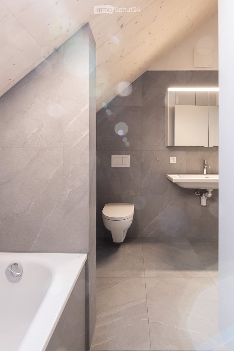 Modern bathroom with grey marble tiles, a bathtub, toilet, sink, and a mirrored cabinet with built-in lighting.