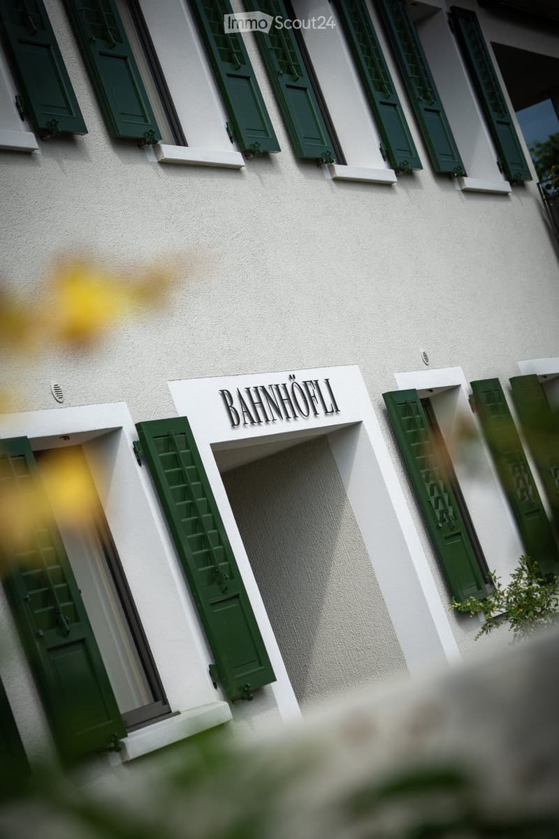 Facade of a building with white walls, green shutters, and a sign reading "BAHNHOFLI".