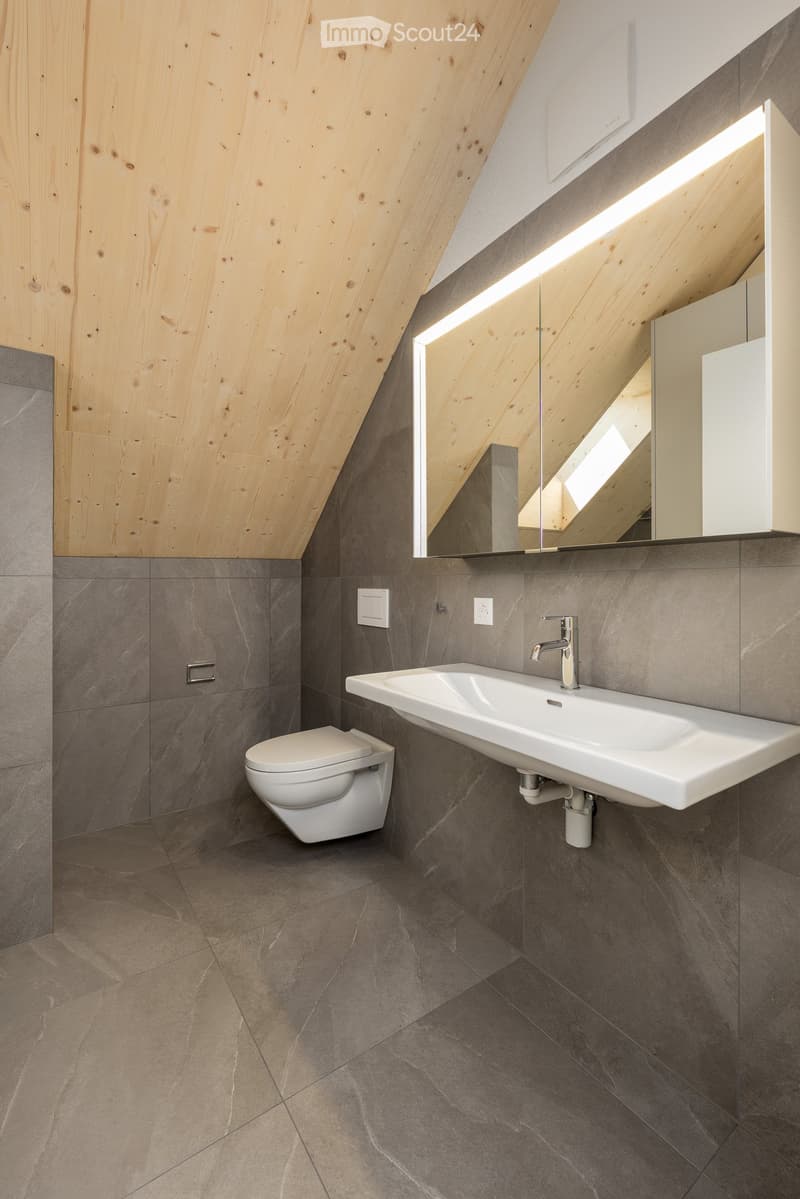 Modern bathroom with grey tiled walls and floor, white toilet, white sink, and a large mirror with built-in lighting.