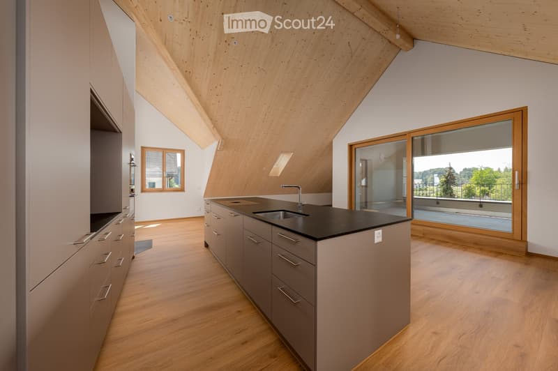 Modern kitchen with wooden ceiling, light wood flooring, grey cabinets, black countertop, and a large window offering a view of the outdoors.