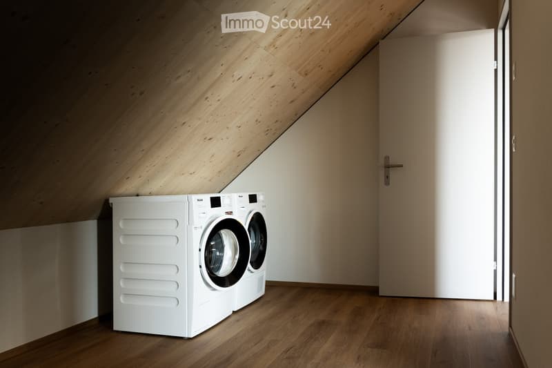 Modern attic space with wooden slanted ceilings and floors, featuring a white washing machine and dryer unit.