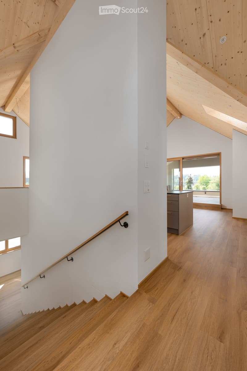 Spacious hallway with wooden flooring, white walls, and a wooden staircase leading upwards. Features modern lighting fixtures and a sleek metal handrail.